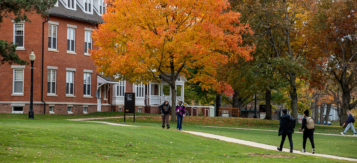 Students walk across campus with Mears Cottage in the background on the campus of Grinnell College. The trees feature the orange and reds of fall foliage. 