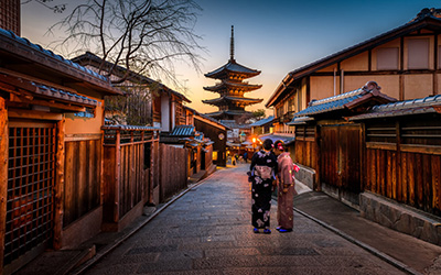 Two women in Kimodos stand in a street in Kyoto with a traditional Japanize temple in the background. 