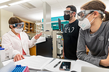 Two students and a professor in eye goggles interact in a chemistry lab. 