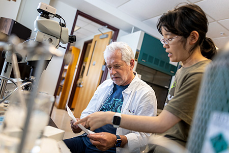 A professor and student compare notes with chemistry equipment surrounding them in a lab.