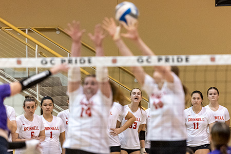 Grinnell Volleyball players cheer a point in the background in focus. In the foreground but blurred, two Grinnell players go for a block.