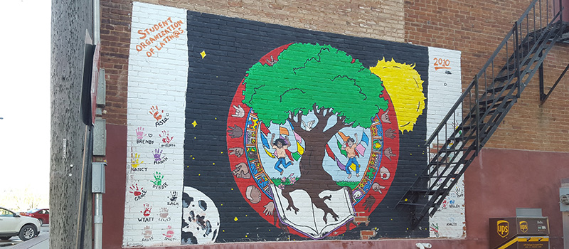 A painted mural featuring a tree, two latinx figures, the sun, and the moon.