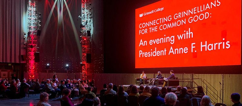 A wide shot of the stage of the Seattle Presidential event President Harris is joined by two alumni on stage. A large projection of white text on red says "An evening with President Anne. F. Harris."