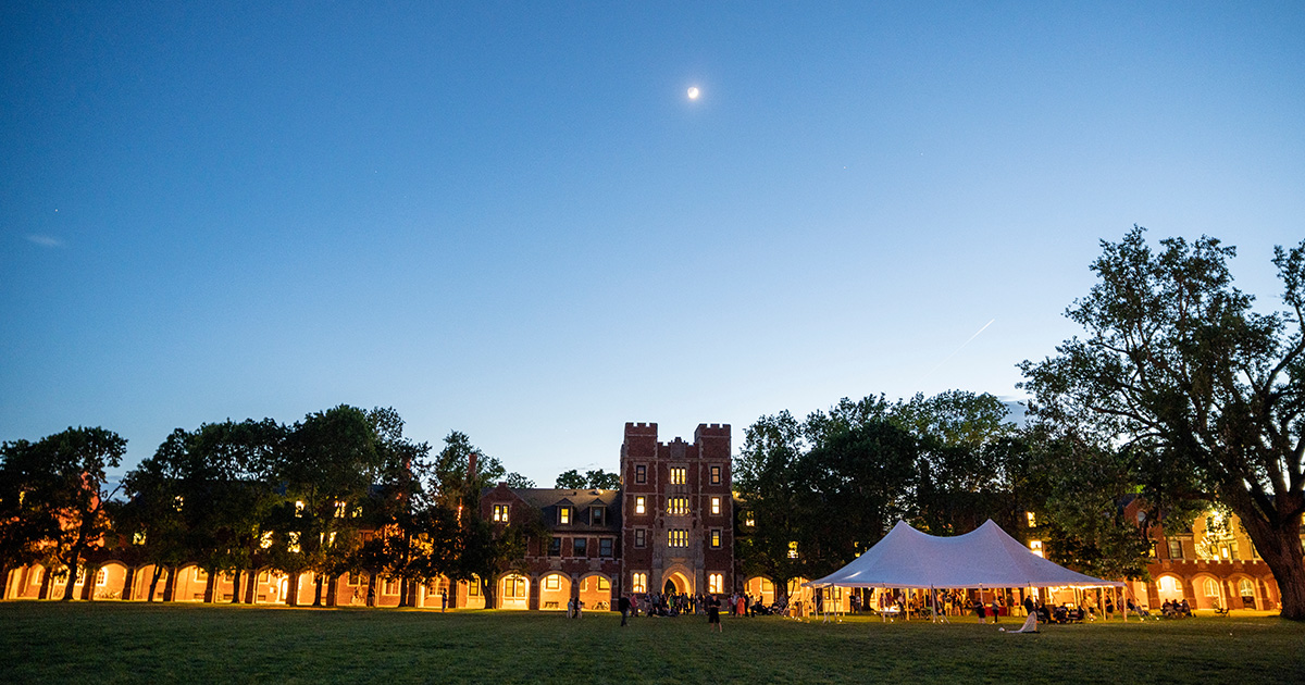 The moon sits over Gates Tower with the loggia illuminated. A tent sits in the foreground on Mac Field.