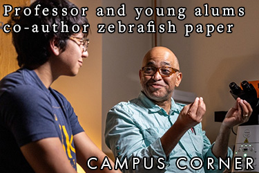 Text: Campus Corner - Professor and young alums co-author zebrafish paper Image: A student and profess chat while microscopes and other equipment are in the background.