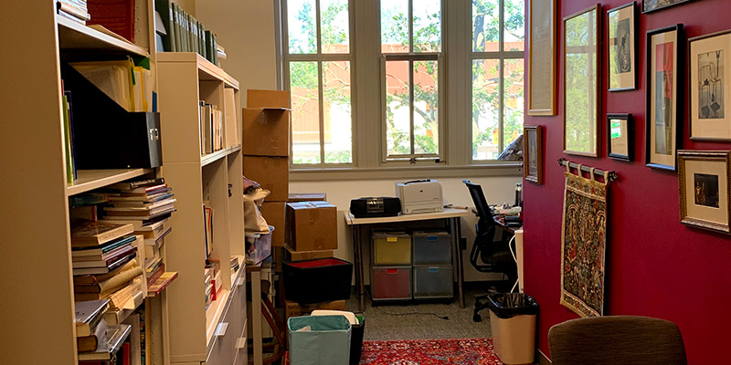 The Harrison Faculty Office. It features bookshelves and red walls covered with framed pictures/images. A desk sits in the back facing windows.