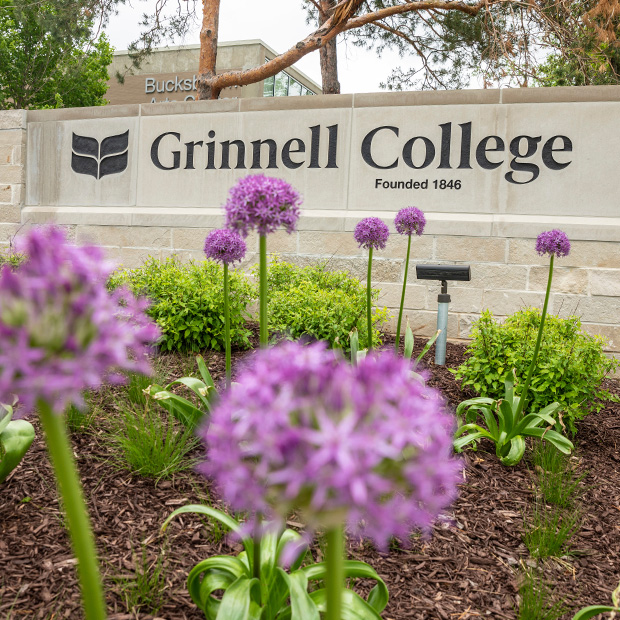 The stone welcome sign to campus. Sign Display: Laurel Leaves logo, Grinnell College. Founded 1846. Purple Flowers are in the foreground. 
